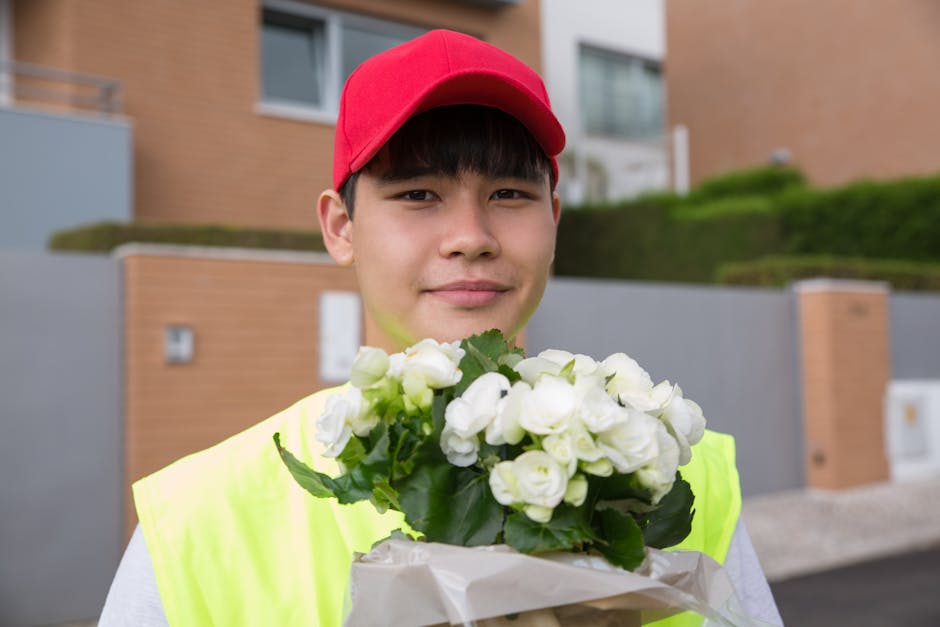 A young male delivery person wearing a red cap and a high-visibility yellow vest is standing outdoors in front of modern residential buildings. He is holding a bouquet of fresh white roses, tightly ar