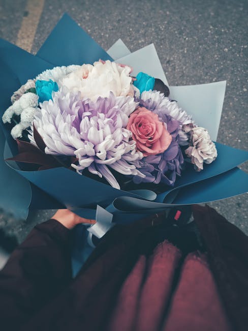 A vibrant floral bouquet featuring large, soft pink roses, purple chrysanthemums, white daisies, and small bright blue flowers. The bouquet is arranged with lush green foliage and wrapped in matte blu