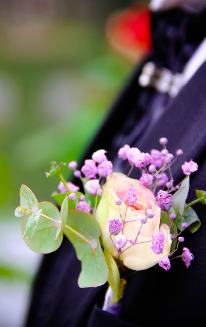 A floral boutonniere featuring a single ivory rose surrounded by delicate purple gypsophila and sprigs of greenery, including eucalyptus leaves with a soft green hue. The arrangement is pinned to a da
