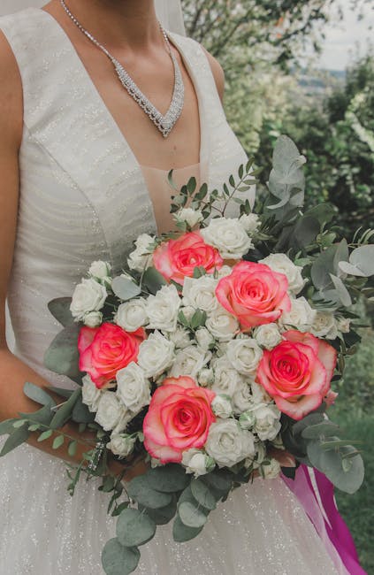 A bride in a sleeveless white wedding gown with subtle glitter accents holds a large floral bouquet outdoors, featuring prominent pink roses surrounded by smaller white roses and silvery eucalyptus le