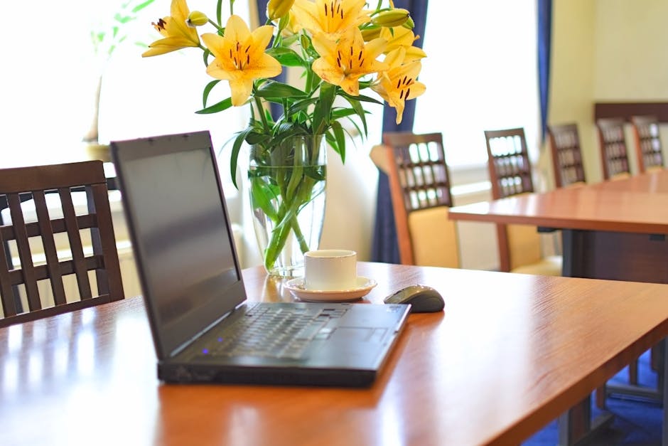 A floral arrangement of vibrant yellow lilies with dark stamens and green leaves is displayed in a clear glass vase, placed on a polished wooden table. The bouquet appears fresh, with open petals show