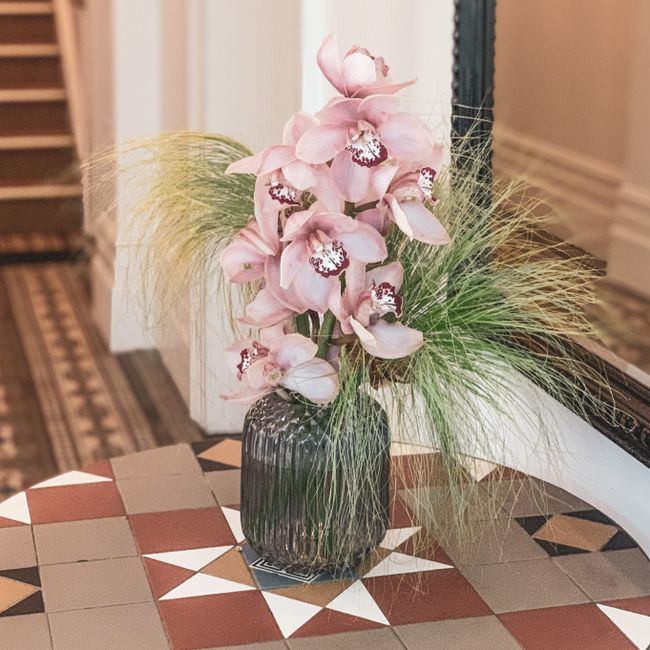 A tall floral arrangement featuring pink orchids with delicate petals and distinctive dark markings on their central lips, set in a dark, ribbed glass vase. The orchids are complemented by wispy, gree