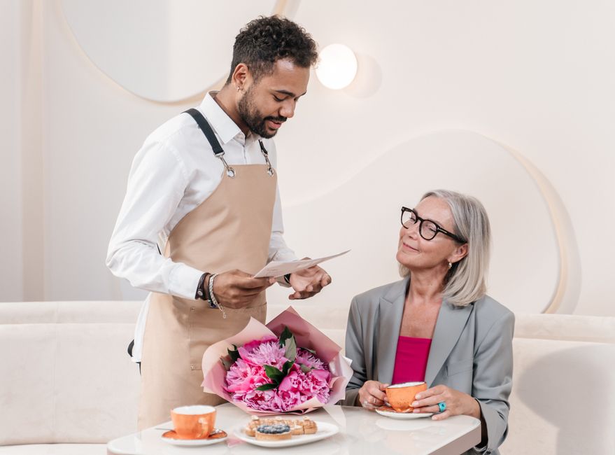 A floral gift of fresh pink peonies and green foliage wrapped in pink paper is being presented by a man in a beige apron and white shirt to an elderly woman with gray hair, glasses, and a gray blazer.