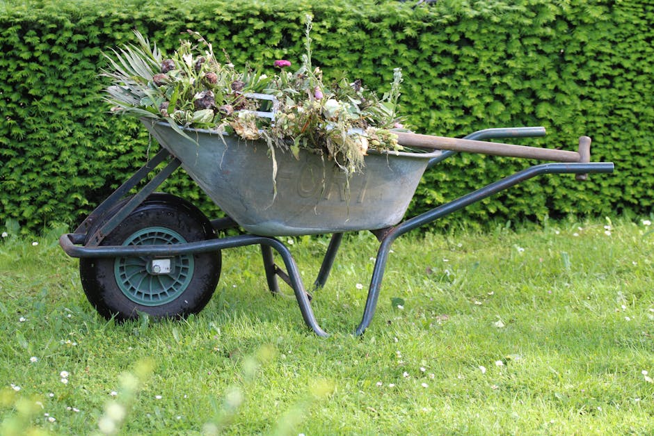 A metal wheelbarrow filled with a mixture of seasonal garden flowers and plant cuttings, including pink, white, and purple blooms, along with green foliage and stems. The wheelbarrow is placed on a gr