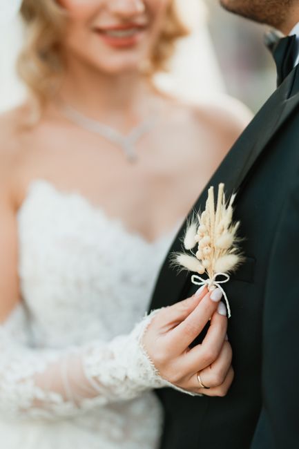 A bride dressed in a lace wedding gown holds a delicate floral boutonniere featuring soft cream-colored pampas grass, small white flowers, and airy plumes attached with a white satin ribbon. She is st