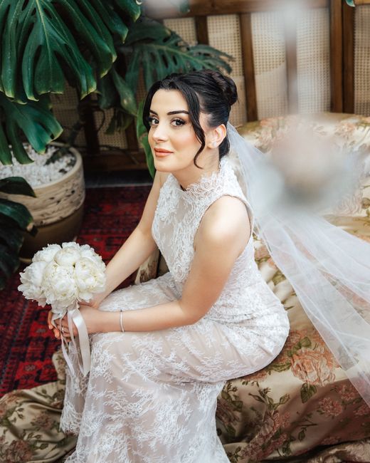 A bride dressed in an elegant white lace wedding gown with a high neckline, sitting on a floral upholstered sofa in a warmly lit indoor setting. She holds a round bouquet of cream-white roses, tied wi