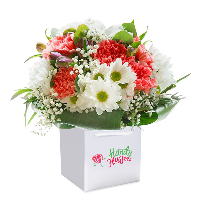 Presented upright in a flawless square white gift bag, this vibrant Chessington bouquet balances soft and bold tones to perfection. Fluffy, coral and peach-hued carnations create a plush foundation, their frilled petals inviting touch and symbolising heartfelt sentiment. Nestled among these are brilliant white daisies with vivid chartreuse centers-bringing an optimistic, sunny lift reminiscent of wild meadows stretching near Chessington's edges. Sprigs of airy white baby's breath weave throughout, softening the arrangement and evoking a sense of lightness, while stray purple-touched buds introduce gentle depth and anticipation. Encircling the entire assembly is a collar of broad, deep green leaves; glossy and substantial, they cradle the blooms like nature's own embrace, echoing the greenery seen in Churchfields and Sir Francis Barker House gardens. The sturdy white ribbon handles and minimalist gift bag subtly signal readiness for gifting-a perfect gesture for a milestone birthday, anniversary, or spontaneous kindness delivered to a friend on Garrison Lane. Captured against a clean, white background, every nuance-from the shimmer of leaves to the pastel gradations in each petal-is highlighted, illustrating both freshness and artistry. This Chessington arrangement celebrates the joy and connectivity that comes from gifting thoughtfully selected, locally arranged flowers.