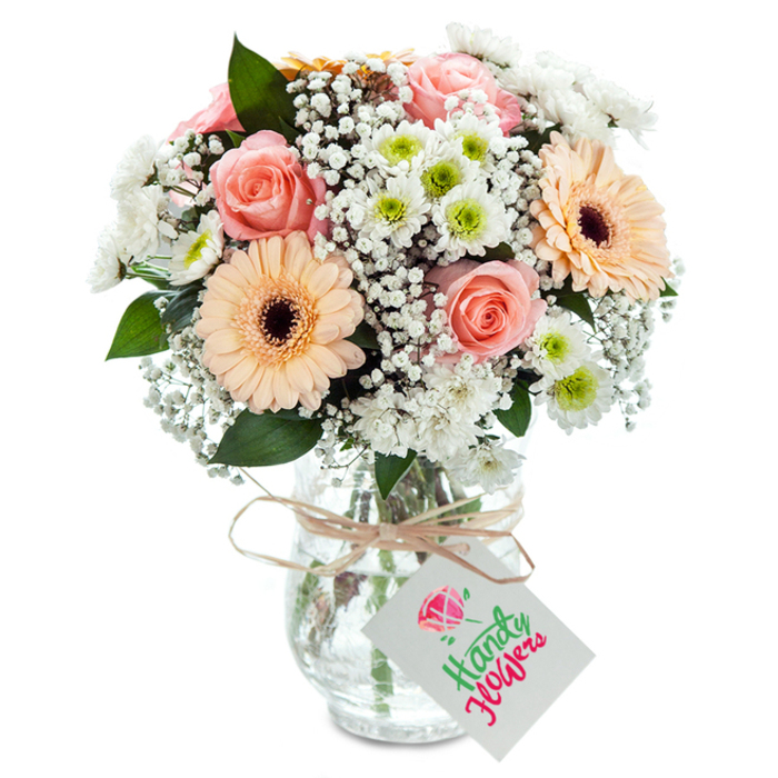 Presented in a beautifully simple clear glass vase, this bouquet is a statement of understated charm and skilled craftsmanship, inspired by Chessington's relaxed, welcoming spirit. Peachy-pink roses with velvety, tightly furled centers form the crown of the arrangement, their classic beauty accentuated by the gentle glow of ambient light. Flanking them are two substantial gerbera daisies in soft peach, their satin petals and distinctive coffee-brown centers drawing the eye and introducing a contemporary playfulness. Spirals of delicate white gypsophila-floating like morning mists along Church Lane-help to soften transitions between the dominant flowers, adding a billowy fullness. Tiny white chrysanthemums, their small greenish-yellow centers glimmering, are dotted throughout, contributing to a textural richness reminiscent of the floral displays often seen in Chessington high street shops. Deep, glossy green foliage, possibly eucalyptus or a similar varietal, nestles among the blossoms, offering contrast and a subtle, refreshing scent. The watery clarity of the glass vase showcases stems expertly arranged for both aesthetics and drink, ensuring lasting vibrancy. Around the neck, a length of rustic twine tied in a neat bow secures a crisp white label-blank and ready for any heartfelt Chessington message you might wish to add. Thoughtfully styled and special in every detail, this bouquet is perfect for commemorating special moments, welcoming a new neighbour, or simply brightening a Chessington home with the promise of renewal and care.