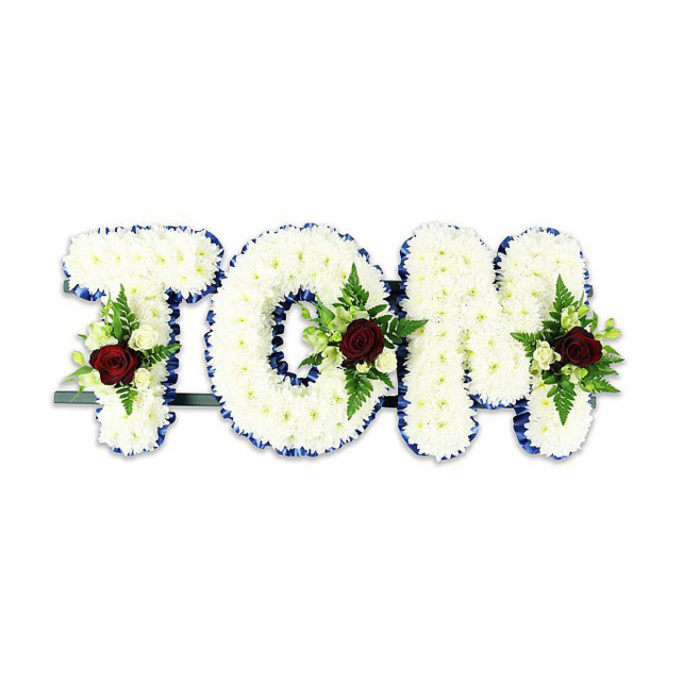 A powerful, personalized floral composition reads TOM in bold, three-dimensional letters, each letter densely composed of tiny, pristine white chrysanthemums that create a soft, tactile surface with subtle shadowing between blooms. The perimeter of every letter is neatly finished with a striking royal blue ribbon that crisply defines each stroke, offering a calm, formal contrast to the snow-white petals. Within each letter a small, symmetrical spray is carefully placed: a single deep, dark red rose stands as the emotional centre, its velvety richness suggesting love and remembrance, while accompanying creamy white spray roses and delicate fern fronds add texture and a green counterpoint. The arrangement's structure is supported by a thin dark green or black metal bar at the base, visible when viewed from certain angles and signalling that this is a standing tribute intended for display at funerals or memorial services. Set against a stark white backdrop, the colours read clearly-white for peace, blue for dignity, red for enduring affection-and the overall effect is both solemn and consoling. There is a gentle botanical scent in a quiet room, the roses giving a faint, classical perfume while the chrysanthemums lend a soft, neutral note. As an experienced local florist I imagine this piece placed in a chapel near Hook or delivered to family homes around Chessington High Street; its measured elegance suits church services, funeral parlours, or a commemorative display at a community gathering. The craftsmanship is understated yet precise, an empathetic floral gesture fashioned to honour memory with grace.