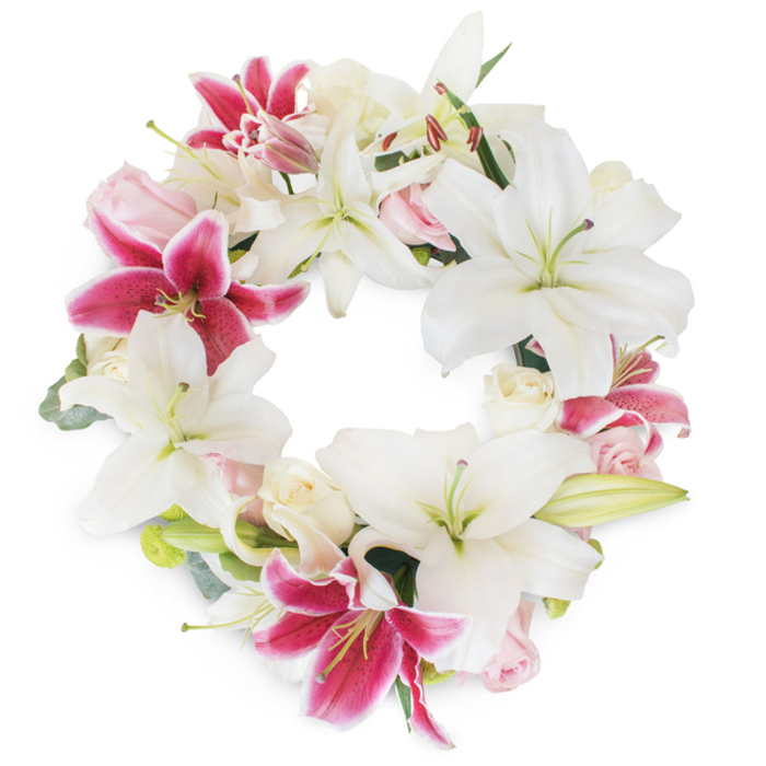 Against a crisp, white canvas, this elegant circular wreath-artfully arranged by skilled Chessington florists-presents an evocative blend of tradition, beauty, and heartfelt remembrance. The composition is dominated by luxurious white Oriental lilies, each bloom opening wide to reveal graceful green pistils and lemon-tipped stamens, their smooth petals curving gently in a gesture of peace and purity. Eye-catching fuchsia pink lilies, with vivid magenta hues and delicate speckles at their outset, are woven expertly between the white blooms, providing rich visual drama much like the bold floral accents seen along Chessington's public parks and avenues. Nestled delicately throughout the circle, soft rose clusters in blush pink and creamy tones add ruffled texture and understated romance; some are compact buds, holding the promise of new life, while others are just beginning to blossom, infusing the arrangement with both hope and nostalgia. Sprigs of muted green foliage, possibly eucalyptus or slender lily leaves, peek through the dense ring, lending a whisper of woodland freshness and subtle structural depth. The open centre and flawlessly symmetrical design evoke a sense of unity and everlasting love-sentiments deeply valued by the Chessington community. This detailed floral tribute, shining brightly against its white background, is especially fitting for commemorations at local venues or as a tender sympathy arrangement delivered to loved ones across Chessington, offering beauty, comfort, and quiet strength.
