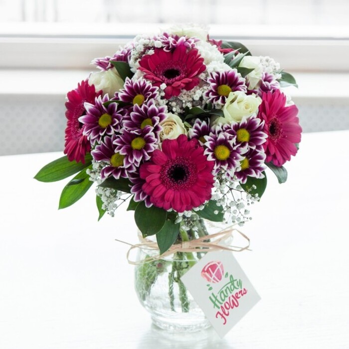 A striking mixed flower bouquet is showcased in a clear, slightly textured glass vase, positioned squarely against a luminous white background that feels like a bright window in a modern Chessington living room. The arrangement is dominated by large gerbera daisies in a saturated fuchsia or magenta, their velvety petals radiating from dark central disks that add intensity and focus to the display. Clustered closely around them are generous, daisy-like chrysanthemums with rich purple tips that gradually fade into creamy white, each bloom finished with a vivid yellow centre that gives the bouquet an almost sunlit glow from within. Between these bolder flowers, creamy white roses are carefully placed, some in full, soft-petalled bloom and others still in elegant bud form, conveying a sense of unfolding moments and quiet romance. Fine, cloudlike sprays of white baby's breath weave through the entire composition, lending a delicate, airy texture that contrasts beautifully with the lush, solid petals of the larger flowers. Deep green foliage frames the blooms, providing structure, height, and a natural contrast that makes the magenta and purple tones appear even more vibrant. The vase itself is squat and jar-like, its neck encircled with rustic twine tied into a relaxed bow, evoking a hand-finished touch often seen in bouquets prepared for same-day delivery across Chessington by local florists like Chessington Flowers. Hanging from the twine is a small, white rectangular tag, decorated with a stylised pink rose emblem and flowing script that hints at a personal message without overwhelming the flowers. Bathed in soft, natural light, every petal appears freshly cut, creating an overall impression of joy, celebration, and thoughtful gifting, ideal for birthdays, thank-you gestures, or simply brightening a hallway in a home near Chessington's green spaces.