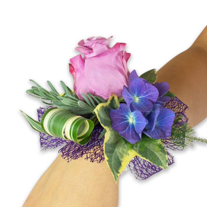 Presented on a light-toned wrist and photographed against a clean white field, this wrist corsage reads like a tiny, wearable bouquet shaped for formal nights and treasured moments. The centrepiece is a large rose with petals that open into a soft gradient of pinks-deeper fuchsia toward the ruffled outer petals, settling to a pale, almost blush core-each petal catching light to reveal a satiny texture. Nestled close are several clusters of rich indigo to purple hydrangea; their miniature florets show faint bluish-white centers that seem to glow slightly against the rose, creating a multi-dimensional, painterly contrast. Architectural foliage supports the flowers: long, silvery-green sprigs (suggesting eucalyptus or dried lavender) add breathy volume and a subtle aroma, while glossy variegated ivy with cream-margined leaves brings a classic, grounding green. A statement element-a wide leaf streaked with white stripes-has been twisted into an elegant curl, introducing a contemporary sculptural accent that plays against the softness of the blossoms. A dark purple mesh with an open weave sits beneath and around the flowers, its texture lifting the composition and reinforcing the hydrangea's deep tones. This corsage feels both refined and youthful, designed for weddings, graduation balls, and prom nights in the Chessington area; I picture it worn for photos in the village or beneath the evening lights near the high street, an accessory that reads as both floral jewellery and a small keepsake of the occasion.