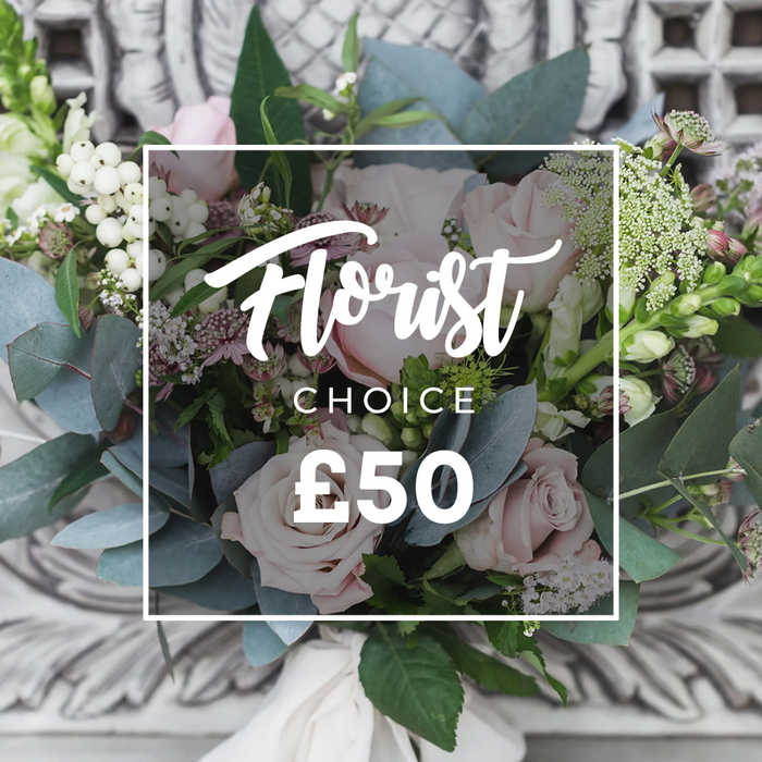 A lush, hand-tied bouquet sits prominently against a softly blurred backdrop of carved, pale stone, the ornate setting lending the composition a refined, almost heirloom quality. At its centre are abundant soft blush roses, their cupped petals layered into romantic rosettes that suggest classic sophistication. Surrounding foliage is generous: dusty blue-green eucalyptus leaves provide a cool, matte counterpoint to the roses, their round silhouettes creating calm negative space. Scattered throughout are clusters of bright white berries-snowberry or hypericum-that add textural contrast and tiny glints of light. Delicate sprays of tiny white filler blooms, perhaps waxflower or astrantia, drift through the design, contributing an airy, meadow-like feel, while smaller deep pink or burgundy blooms give pockets of shadowed warmth. Taller green-and-white spike flowers peek at the edges, offering vertical rhythm and a hint of movement. The bouquet is tied with a cream fabric ribbon visible at the stem base, reinforcing the hand-finished, bespoke nature of the piece. Over the image a clean white frame carries copy identifying the arrangement as a Florist choice with a clear price point-text that sits purposefully but unobtrusively over the floral scene. Lit in soft daylight, the overall impression is one of curated restraint and fresh craftsmanship. This is the kind of arrangement Chessington Flowers would prepare for a special occasion or to grace a Georgian mantelpiece in Kingston, reflecting local floristry skill and seasonal freshness without fuss.