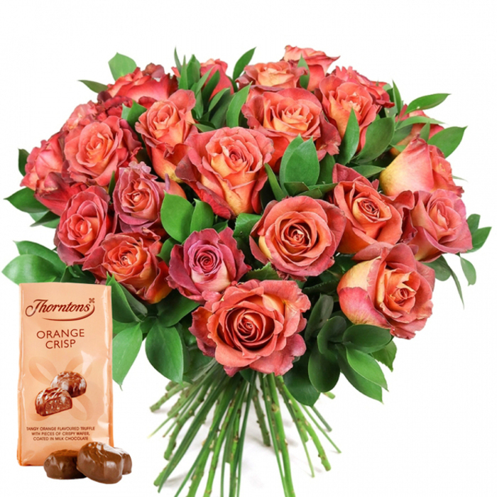 The image presents a striking bouquet overflowing with plush roses - their petals unfurling in every shade from creamy peach to vibrant coral and bold, deep pink, with touches of yellow at the centers lending dimension and a hand-painted quality. The arrangement sits boldly against a white background, allowing every warm hue and velvety texture to stand out. Glossy, dark green foliage, likely including ruscus or eucalyptus, surrounds the roses, creating a sense of balance and organic fullness much admired by Chessington's discerning flower lovers. In the foreground, the classic pairing of indulgent Thorntons Orange Crisp chocolates is artfully presented: a rectangular, ivory-hued pouch upright beside two milk chocolate truffles, their surfaces catching the light and hinting at a creamy, zesty orange center with bits of crisp wafer. The bouquet's stems are elegantly visible at the base, emphasizing its fresh, local preparation - the kind you'd expect from Chessington Flowers just steps from the town centre. The ensemble evokes joyful celebration - perfect for birthdays, Valentine's Day, motherly appreciation, or any occasion deserving of something special. The thoughtful curation reflects the skill and heart that Chessington's finest florists bring to every meaningful gift.