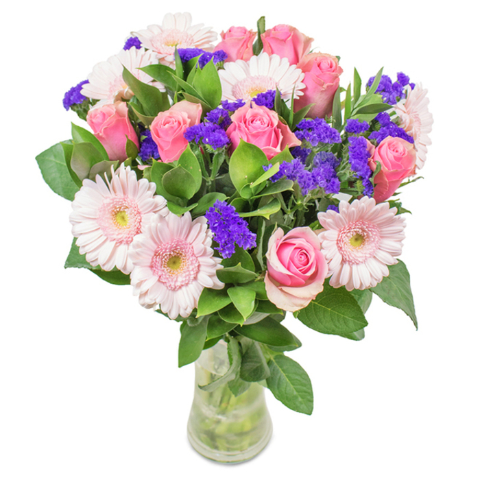 An elegantly composed bouquet, hand-arranged in Chessington and displayed in a pristine clear glass vase, rests against a crisp white backdrop. At its heart, roughly 7-9 soft-to-medium pink roses exhibit the classic spiral shape, some petals tightly furled with promise, others just beginning to stretch open-each bloom echoing a sense of new beginnings akin to those seen each spring in Chessington's parks. These roses are complemented perfectly by 5-7 pale pink Gerbera daisies, their expansive, sun-shaped heads lending brightness, each centre fading to near-white and radiating a warm, welcoming light. Clustered between these focal blooms are vivacious purple statice, their feathery textures and deep hues infusing zest and intrigue, much as local wildflowers enliven the paths at Chessington Wood. Deep green foliage encircles the bouquet, adding abundant volume and a sensation of freshly gathered abundance. Through the glass vase, stems and clean water gleam with promise, suggesting both meticulous freshness and the immediate delight of Chessington's dependable flower delivery. Designed to enchant for birthdays, anniversaries, Mother's Day, or as a heartfelt gift to cheer a home or office in Chessington, this arrangement radiates joy, celebration, and sincere affection-an enduring reflection of local expertise and care.