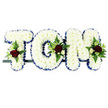 A bespoke funeral name tribute in the photograph spells out TOM in large block capitals, the three letters aligned in a neat row on a slim dark frame as if ready to rest on a hearse or at the front of a memorial service. Each letter is completely covered in tightly packed white chrysanthemums, their small petalled blooms overlapping to create a smooth, cushion-like surface that feels calming and serene. Surrounding the outside edge of every letter, a royal blue satin ribbon forms a crisp pleated border, adding both definition and an air of traditional dignity often favoured for services in and around Chessington. Carefully placed on the 'T', 'O', and 'M' are small, eye-catching clusters of deep red roses, their velvety petals half-open to show rich crimson centres. They sit alongside petite white or cream spray roses and slender white flowers reminiscent of freesia, all supported by soft green fern fronds and glossy foliage that spill gently over the chrysanthemum base. The white background of the image keeps the focus entirely on the tribute, highlighting the contrast between the snowy flowers, the strong blue ribbon, and the rich reds and greens of the accent sprays. It is the kind of personalised arrangement an experienced florist in Chessington Flowers might create for a family wishing to honour Tom by name, sending a message of purity, love, and remembrance. The design feels both traditional and deeply personal, offering a focal point of comfort at a time of grief for families across Chessington, Hook, and the nearby Surrey area.