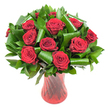 A classic, rounded bouquet of a dozen or more deep crimson roses sits perfectly poised in a tall, slender ruby-red glass vase. Each rose is fully opened, its velvety petals spiraling from a tight centre into broad, soft faces that catch the light and read as almost wine-dark at the petal edges. The blooms are nested in a generous spray of dark green foliage - glossy Ruscus- or Salal-like leaves form a dense, verdant backdrop while larger, bright emerald leaves are expertly rolled and folded into architectural loops and cylinders that cradle individual heads and add a modern sculptural line to the composition. The vase has a subtle ribbed or twisted texture that creates vertical highlights and soft reflections, the translucent red glass deepening the rose colour and anchoring the arrangement with elegant weight. Soft, even studio lighting isolates the bouquet against a clean white background, sharpening the contrast so the red petals and lush greens pop with clarity and depth. Close your eyes and you can almost sense the faint, classic scent of roses, the cool smoothness of glass under your palm, and the tactile contrast between satiny petals and glossy leaves. This arrangement reads as refined romance - ideal for an anniversary or Valentine's gesture, or as a luxurious table centrepiece for an intimate celebration. A local touch: this style is perfect for delivery across Chessington and nearby Tolworth or Kingston, bringing a neighbourhood florist's care and seasonal freshness to every heartfelt occasion.