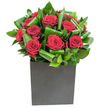 In this elegantly modern composition, deep crimson roses take centre stage, each bloom unfurling in tight, velvety spirals that catch the light like small, soft beacons of passion. The roses are nestling within broad, glossy emerald leaves that have been expertly curled and intertwined to form a rhythmic framework, the foliage creating overlapping arcs and pockets of shadow that give the cluster sculptural movement and visual depth. The arrangement sits in a crisp, matte black cube - its square lines grounding the composition with an urbane, architectural presence that contrasts the roses' plush texture. Bright, steady studio lighting glances off the sheen of the leaves and brings out the rose petals' velvet sheen, making the crimson tones read both warm and intense against the coolness of the vessel. Up close you can almost sense the tactile contrast: the soft, yielding petals against the taut, slightly waxy surfaces of the greenery; the matte vessel's understated grain beneath your palm. A gentle, clean fragrance seems to rise from the bouquet - classic rose lifted by the green, almost citrus-clean notes of the leaves - a scent that feels refreshing rather than heavy. As a local florist, I imagine this piece placed on a contemporary coffee table in a Chessington home or at the corner of a glass-topped desk near Chessington High Street, an arrangement that speaks of considered romance and composed drama. It is suited to anniversaries or a heartfelt expression of admiration, a design that feels intimate but decisively modern and memorable.